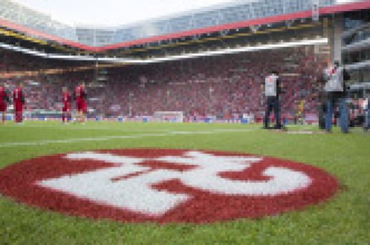 © view - die agentur Aufnahme im Inneren des Stadions. Man sieht im Vordergrund das FCK-Logo auf dem Rasen, im Anschluss das Spielfeld mit Spielern des 1. FCK und im Hintergrund die Westtribüne mit Zuschauern. © view - die agentur