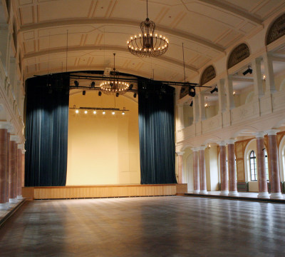 Durch die halbrunden Fenster dringt Licht von außen in den großen Saal der Fruchthalle. Ein schwerer Vorhang ist halb geöffnet und gibt den Mittelteil der Bühne frei. Obwohl der Saal leer ist wirkt er durch den balkonartigen Aufbau und die schlichten, marmorierten Säulen sehr festlich.