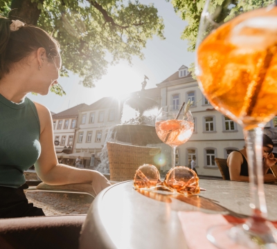 Eine junge Frau sitzt an einem Tisch mit zwei Getränken. Im Gegenlicht sieht man den Martinsbrunnen im Hintergrund.