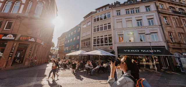 Man sieht den Platz vor der Stiftskirche im Sommer. Im Vordergrund eine Frau, die ein Gebäude fotografiert. Im Hintergrund Außengastronomie.